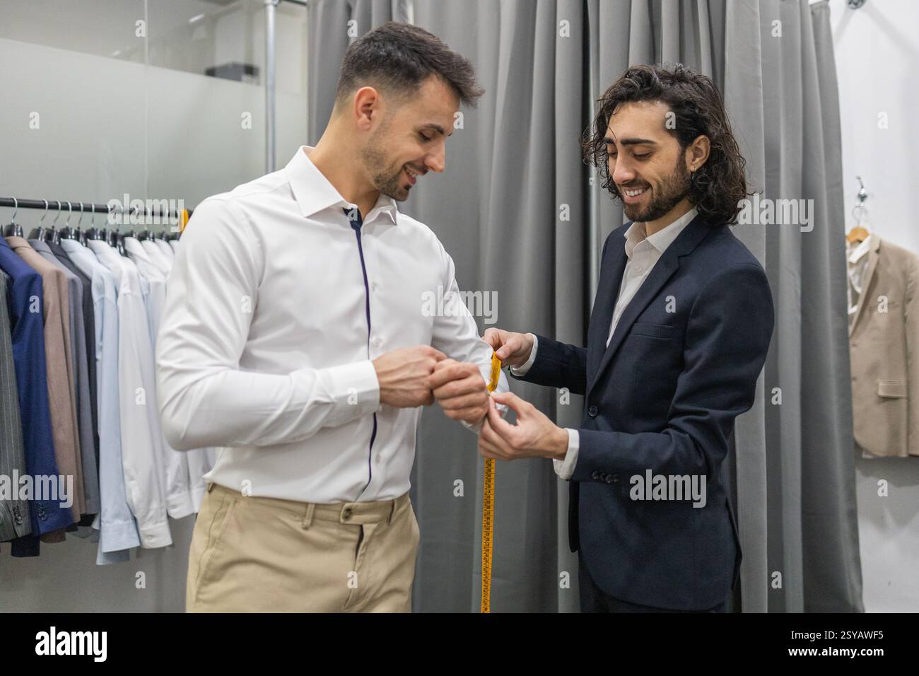 Un sarto misura un cliente per una tuta personalizzata in un negozio di tute. La scena cattura precisione e professionalità nella sartoria, con una gamma di abiti Foto Stock
