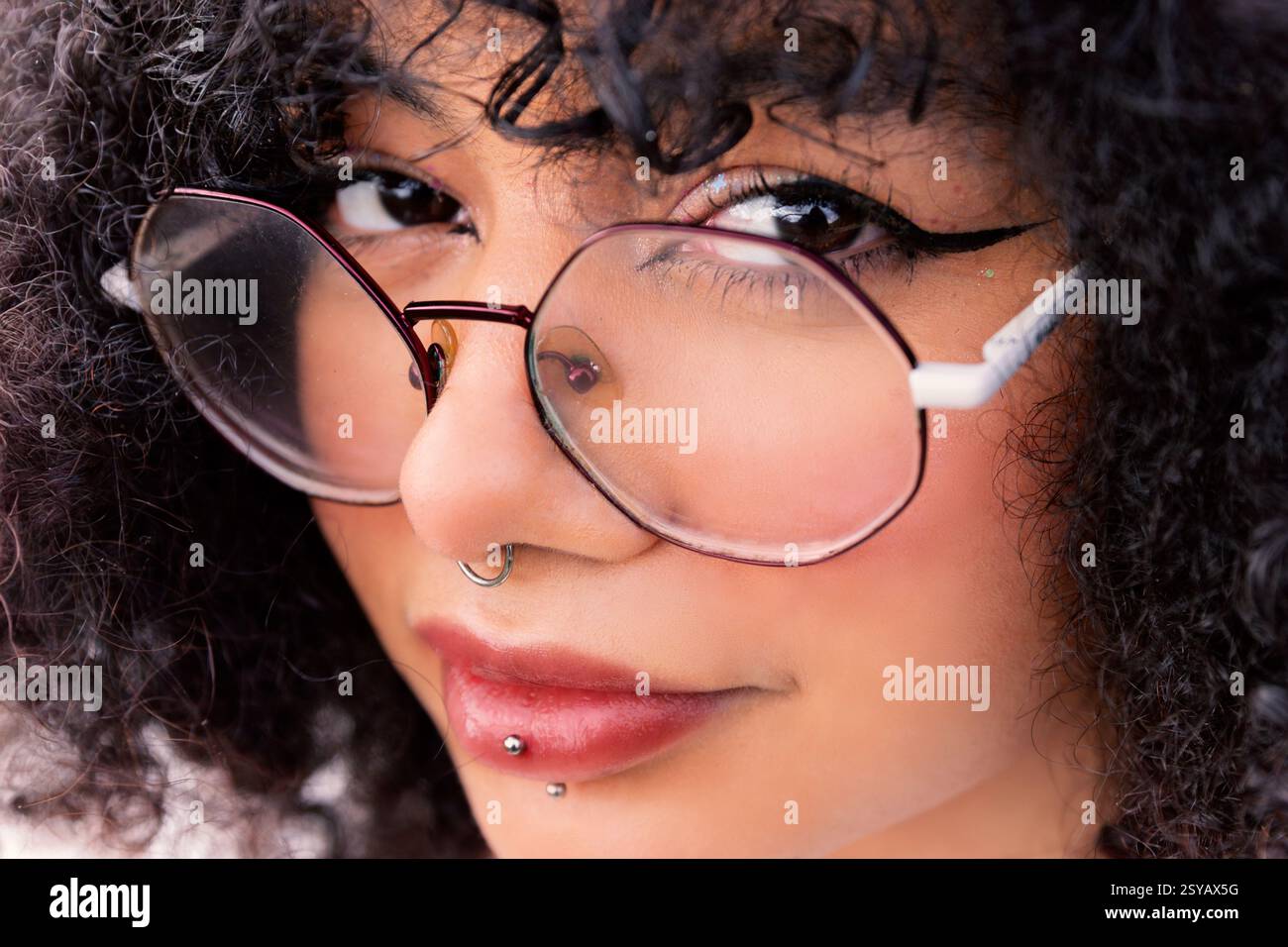 Primo piano di un'elegante donna latina con grandi occhiali, capelli ricci e piercing facciali, che sfoggiano un look espressivo e sicuro. Cattura il moderno b Foto Stock