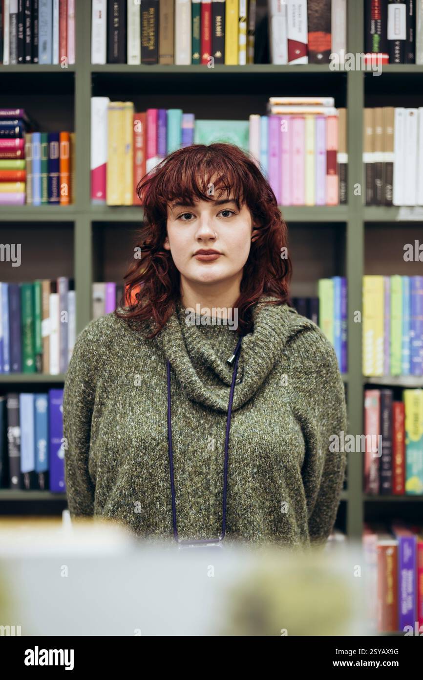 Una giovane donna accademica fa una pausa ponderata in una biblioteca, circondata da librerie colorate piene di numerosi libri, mostrando un ex contemplativo Foto Stock