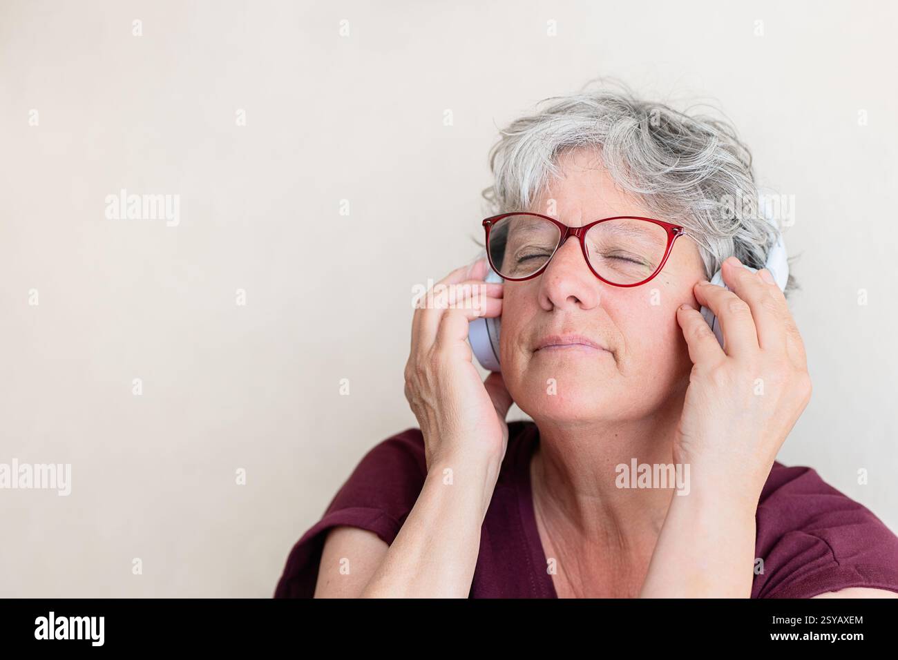 Una donna anziana con capelli e occhiali grigi ama ascoltare musica attraverso le cuffie, gli occhi chiusi, creando un'atmosfera tranquilla. Indossa una camicia rossa scura e un bagno Foto Stock