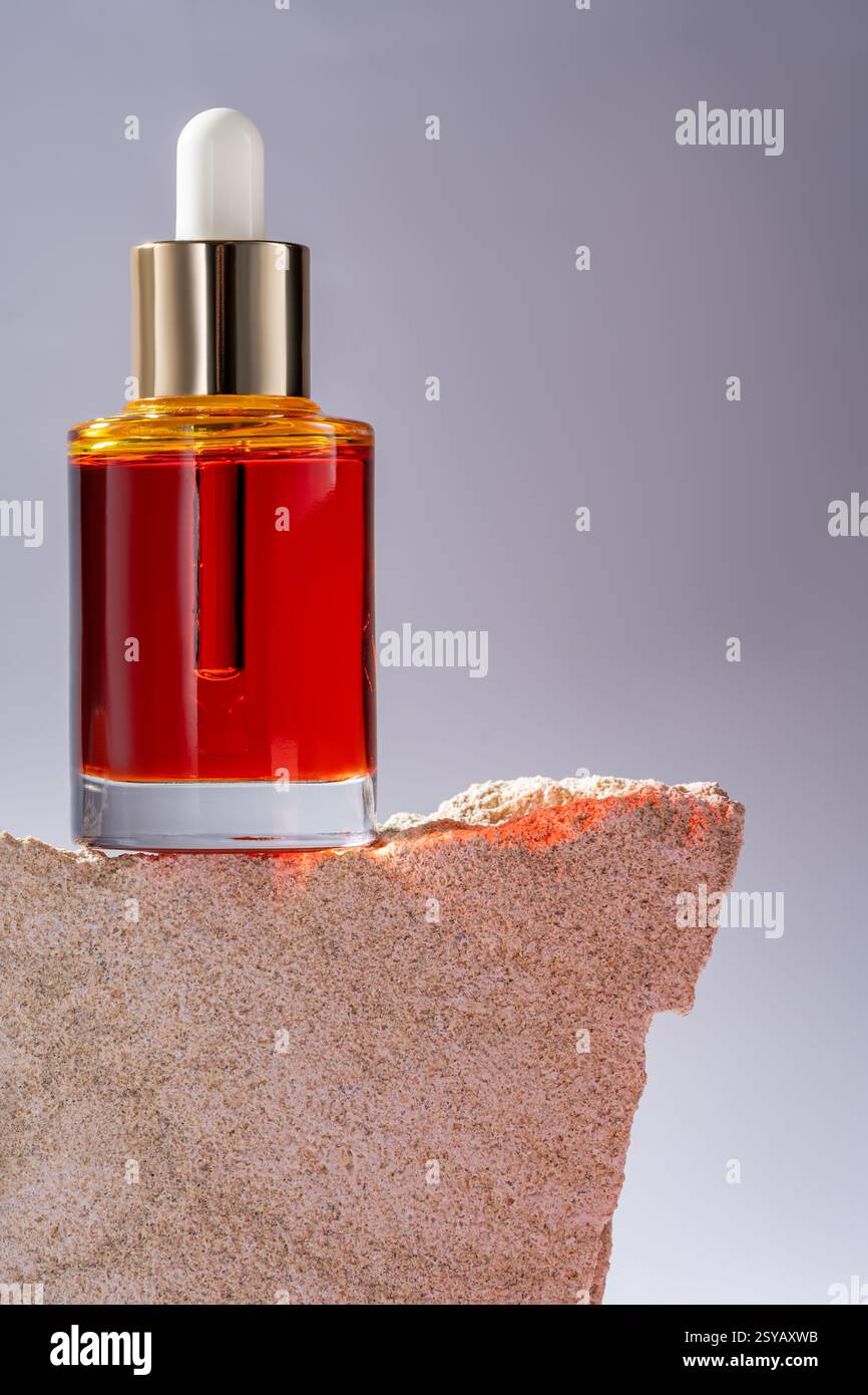 Siero cosmetico rosso vivace in una bottiglia di vetro trasparente con contagocce, elegantemente esposto su un robusto podio in pietra. Foto Stock