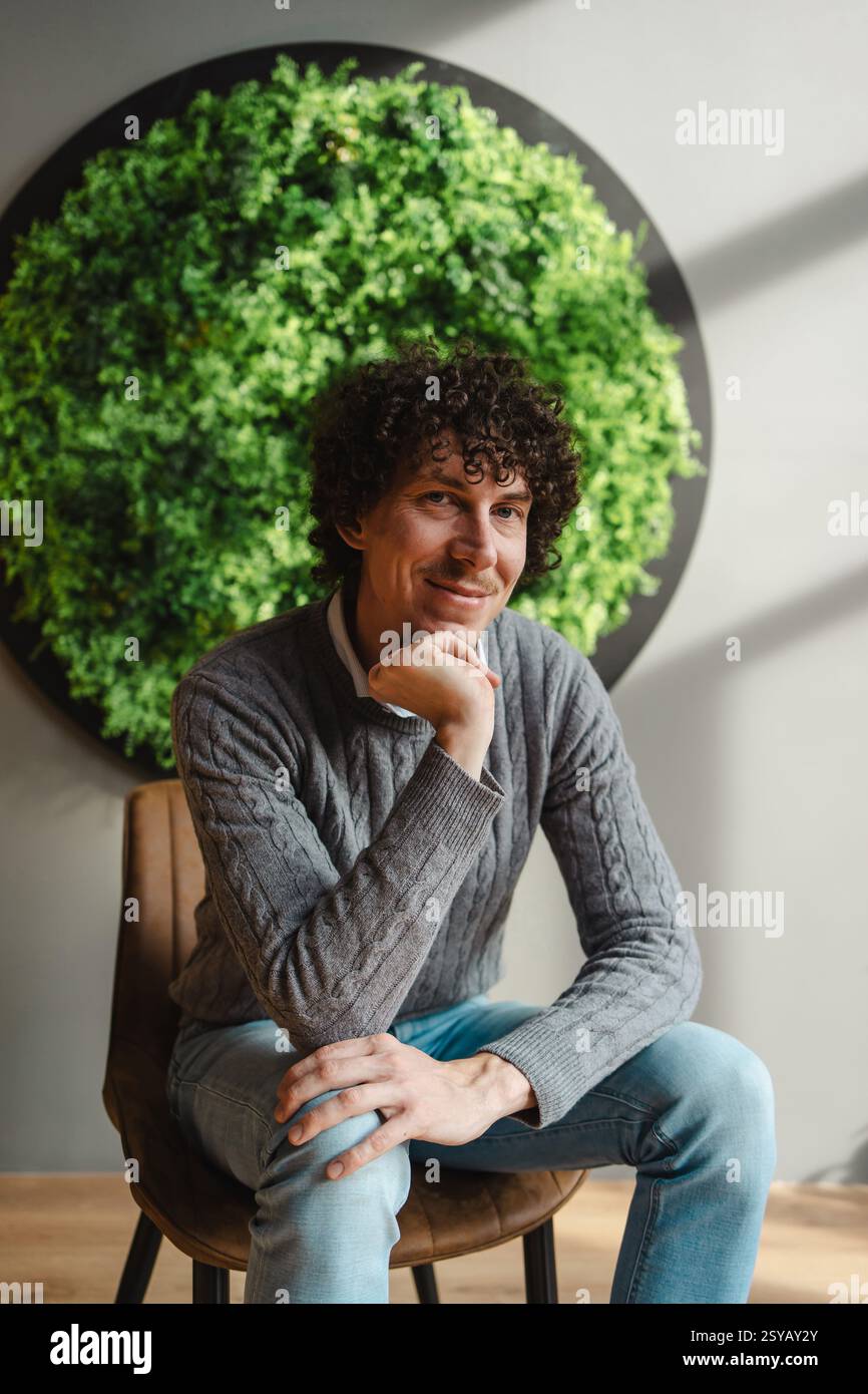 Un giovane uomo con i capelli ricci seduto con cura, incorniciato da una lussureggiante parete verde. Questa immagine mette in risalto i temi dell'ecologia e dello sviluppo sostenibile in Am Foto Stock