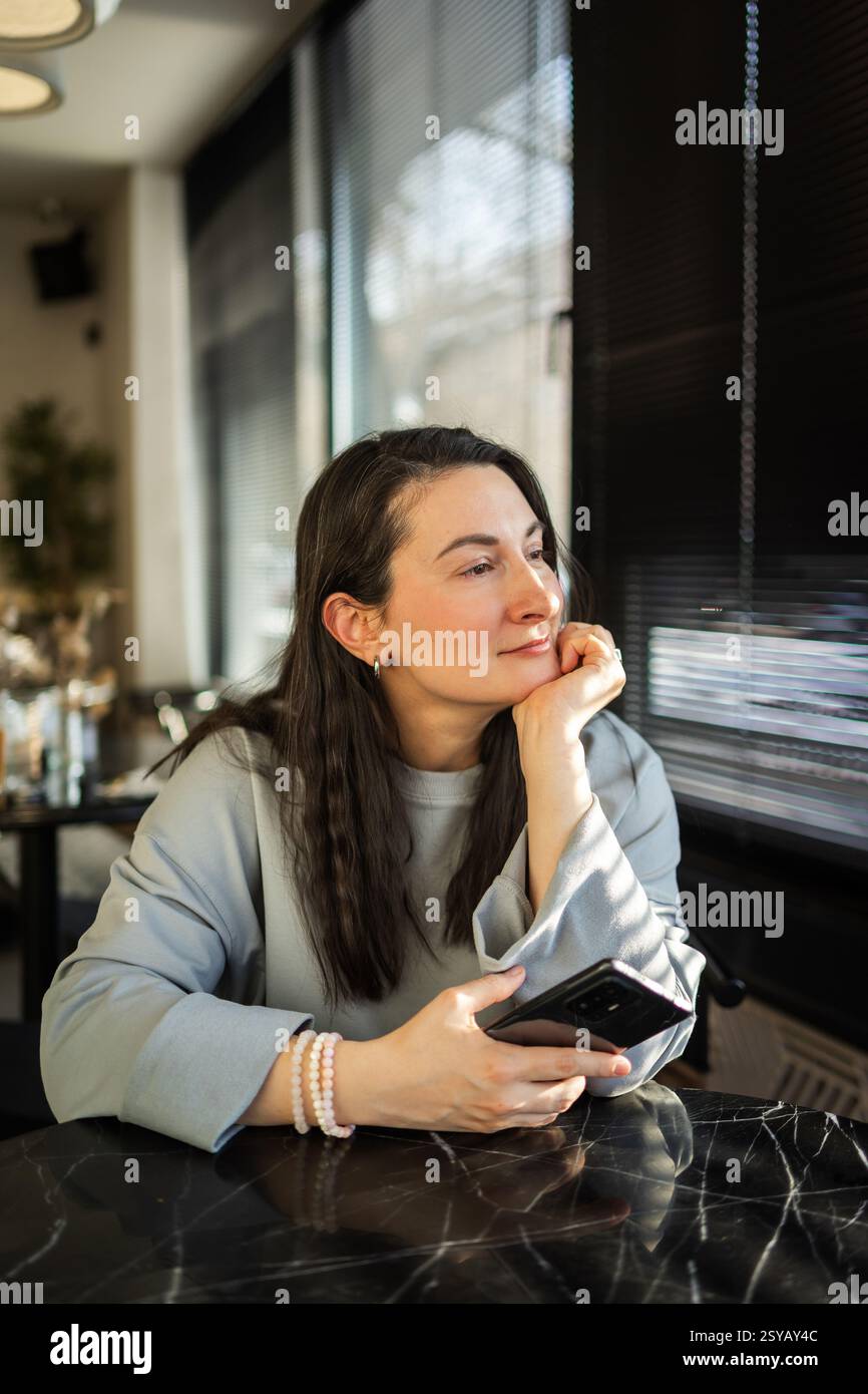 Una donna di mezza età siede pacificamente in un bar, guardando fuori mentre tiene in mano uno smartphone. La luce del sole mette in risalto la sua espressione premurosa Foto Stock