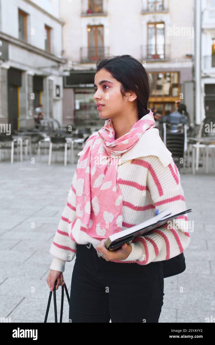 Una studentessa araba si trova in una strada della città con un notebook e una borsa tote. Indossa un maglione a righe rosa e bianco e una sciarpa rosa. Backgroun Foto Stock