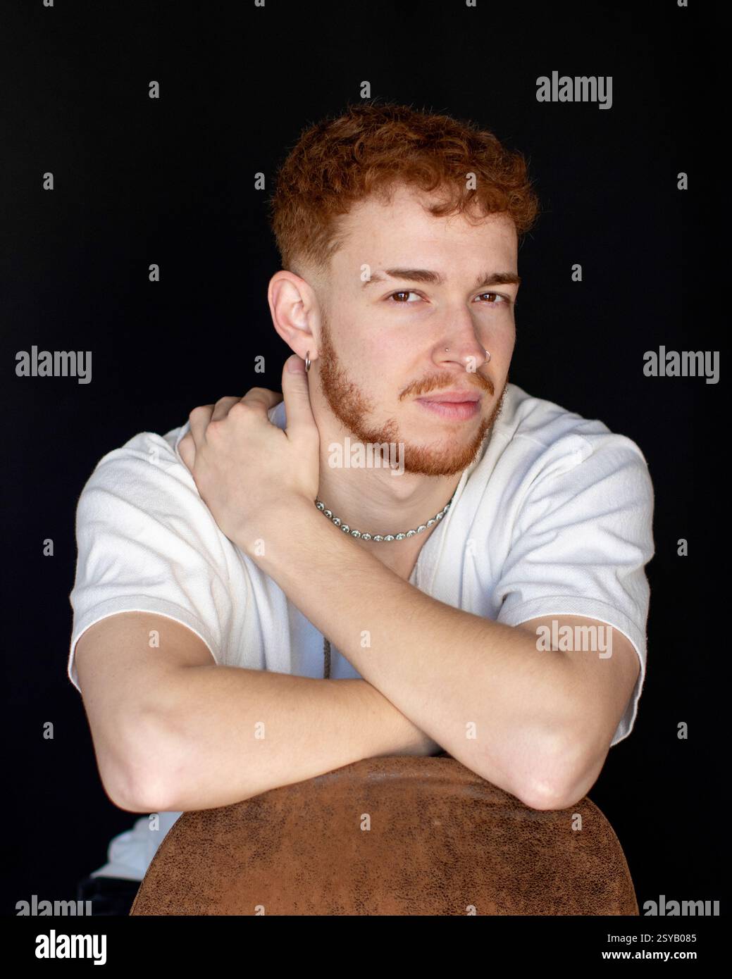 Ritratto di un giovane uomo con i capelli rossi e un'espressione premurosa, seduto su uno sfondo nero. Indossa una camicia bianca e una collana d'argento, annuncio Foto Stock