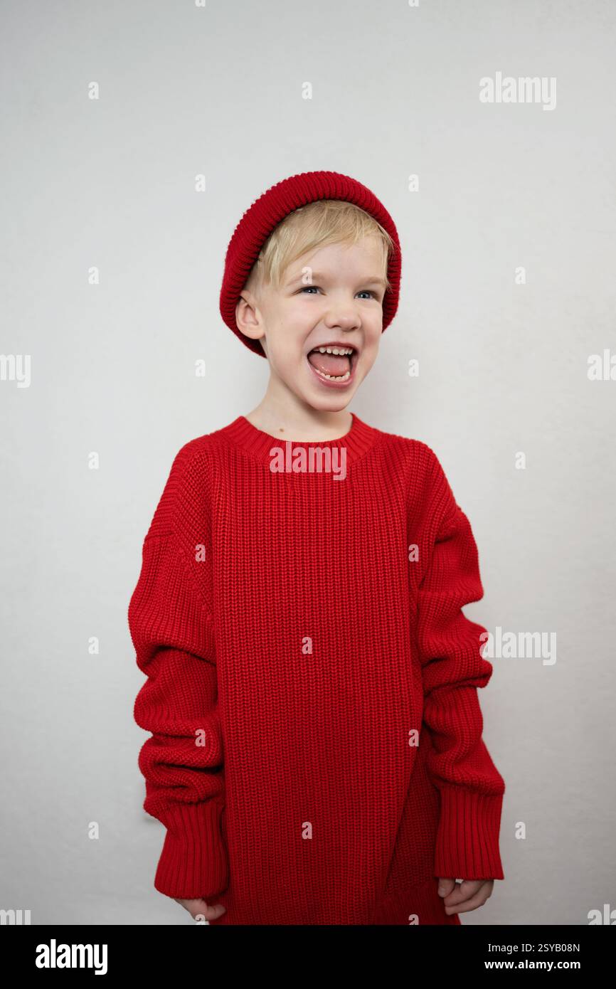Un bambino allegro con un comodo maglione rosso e un berretto abbinato ride gioiosamente su uno sfondo semplice. L'immagine cattura un momento di felicità spensierata Foto Stock