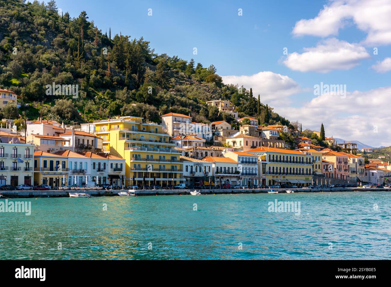 Vista pittoresca delle colorate case sul lungomare di Gythio e degli affascinanti edifici lungo la costa, con lussureggianti colline verdi sullo sfondo sotto una luce luminosa Foto Stock