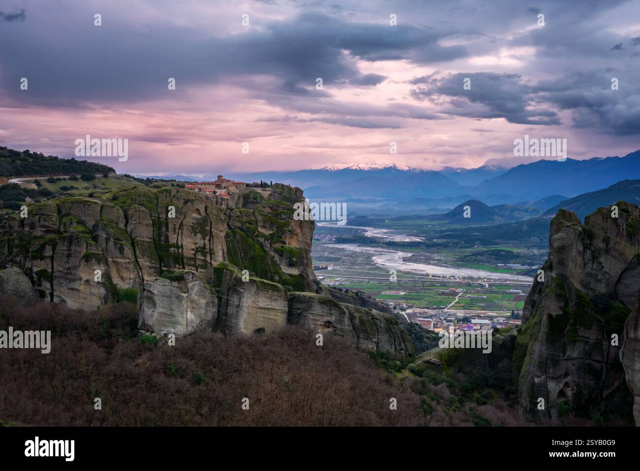 Accattivante vista al tramonto dello storico monastero della Santissima Trinità arroccato in cima a torreggianti scogliere in Grecia, con una valle serena e un lontano pe innevato Foto Stock