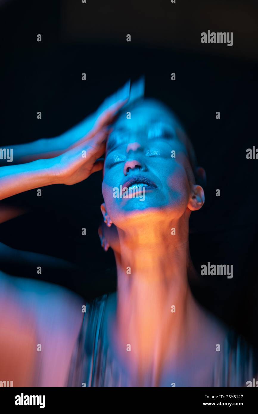 Un suggestivo ritratto di una donna calva, catturato con una vivace luce blu e arancione. La sua posa espressiva trasmette un senso di movimento ed emozione, enhanc Foto Stock