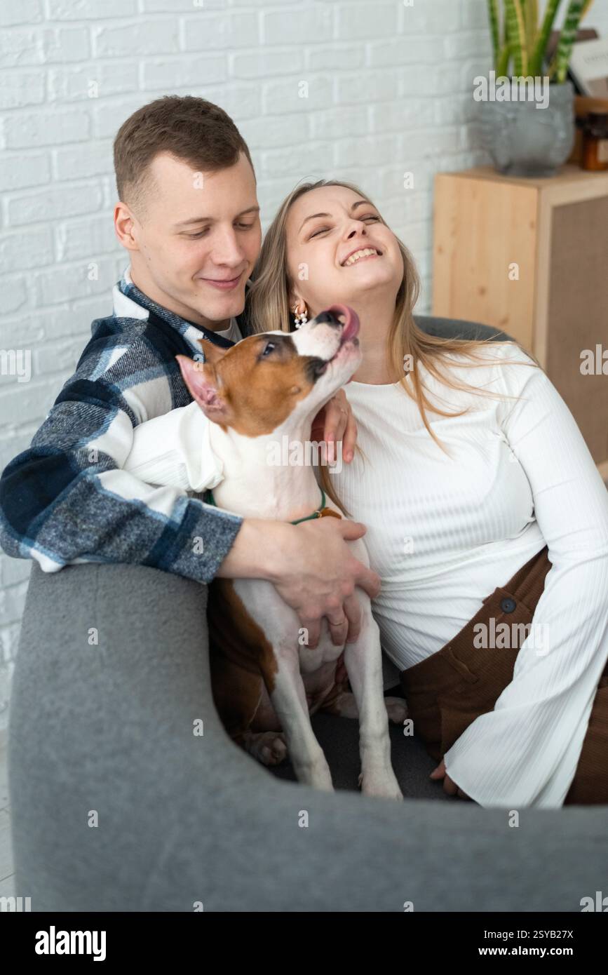Un uomo e una donna gioiosi condividono un momento di amore con il loro cane bianco e marrone giocoso, seduto comodamente nella loro casa elegante e accogliente. Foto Stock