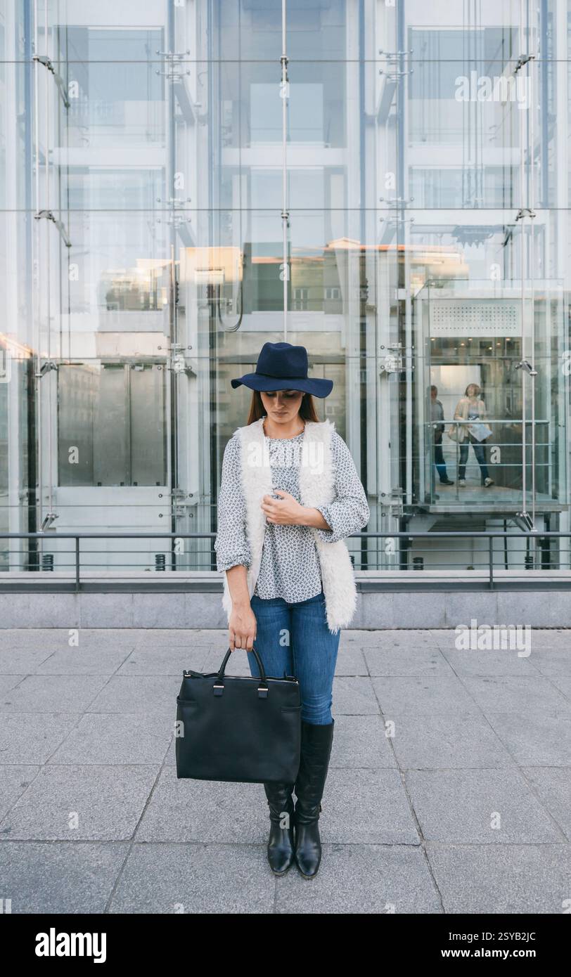 Una donna alla moda si erge di fronte a un moderno edificio in vetro con un cappello a teglia larga, jeans blu e un gilet bianco. Ha una grossa mano nera Foto Stock
