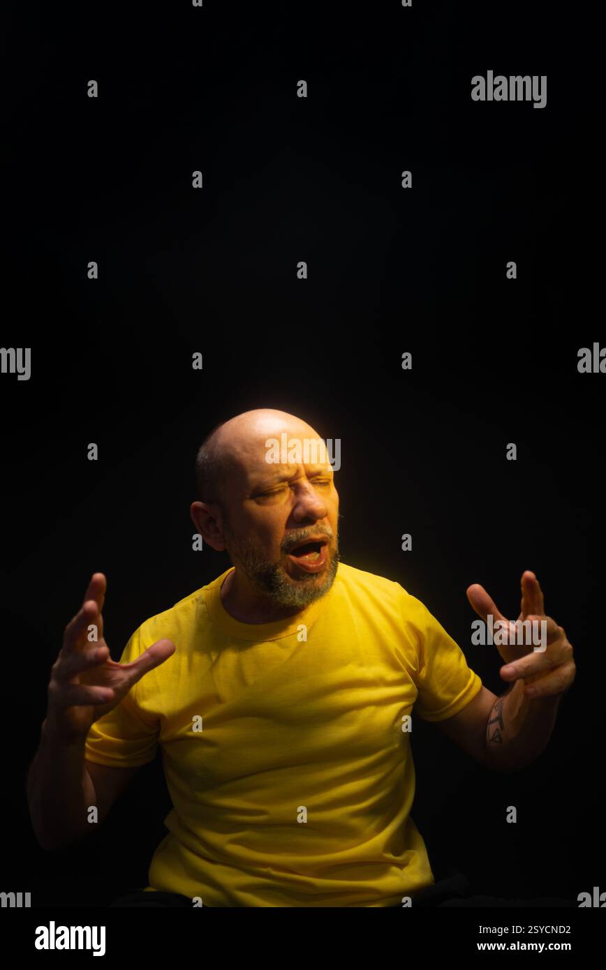 Uomo maturo, anni '50, calvo e barbuto, indossa una camicia gialla, fa gesti delle mani e mostra preoccupazione, paura o disagio. Concetto di assistenza psicologica. Bra Foto Stock
