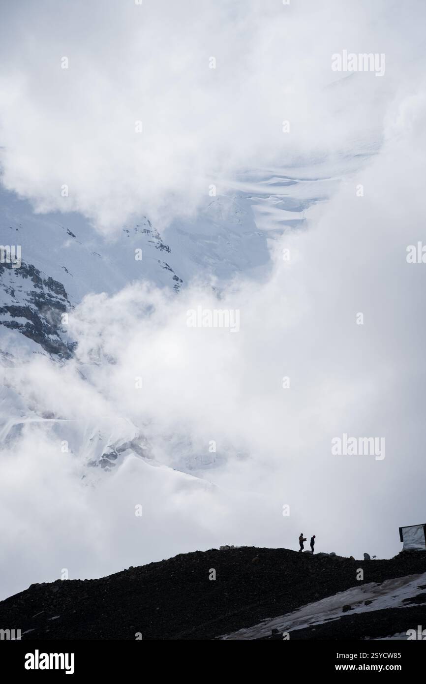 Tiro verticale di due uomini che lavorano sotto enormi montagne innevate avvolte da nuvole, Pamir, Kirghizistan Foto Stock
