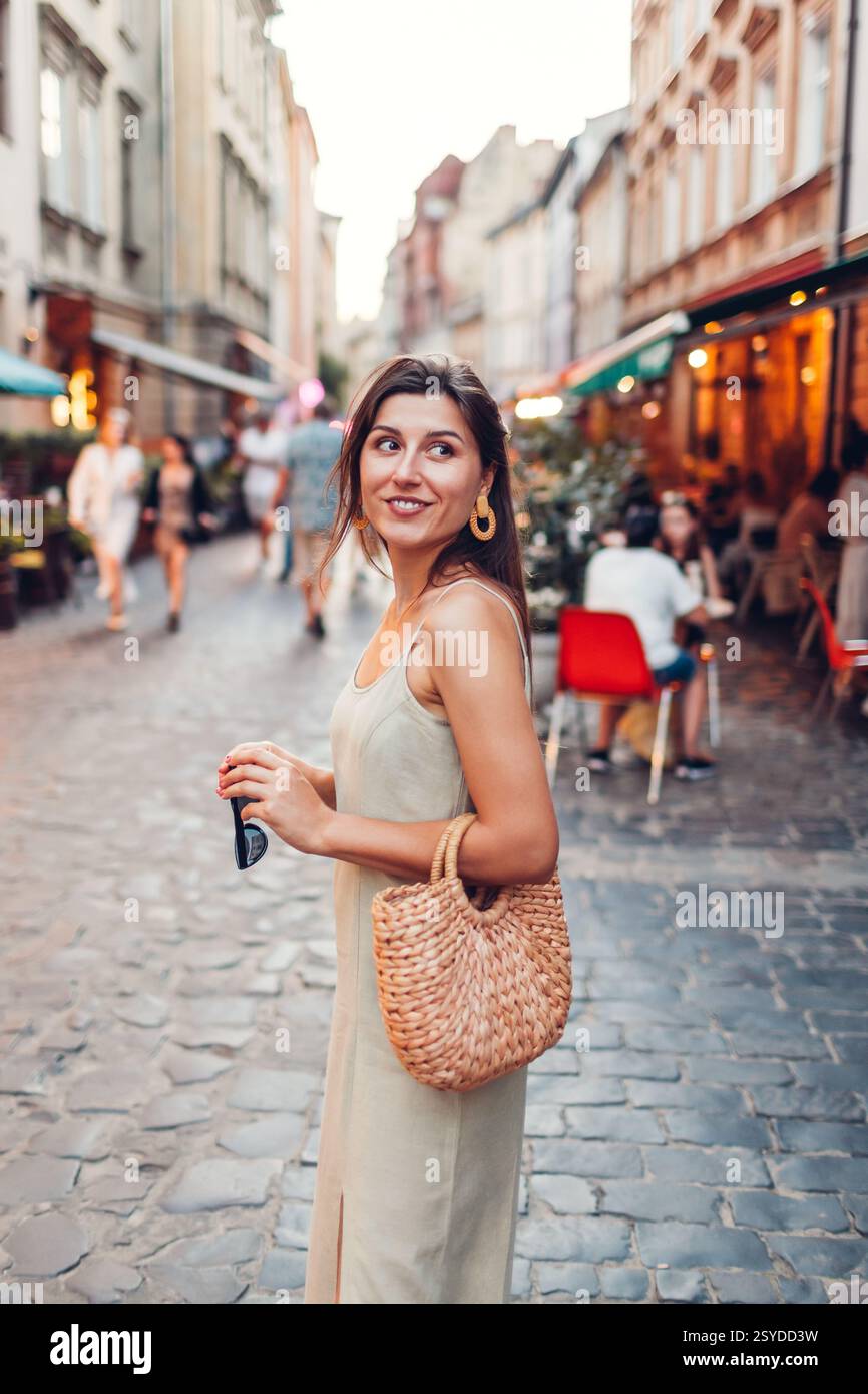 Ritratto di una giovane donna alla moda che indossa l'abito estivo in città per strada accanto al ristorante la sera. Elegante borsa in paglia. Accessori femminili e panno Foto Stock
