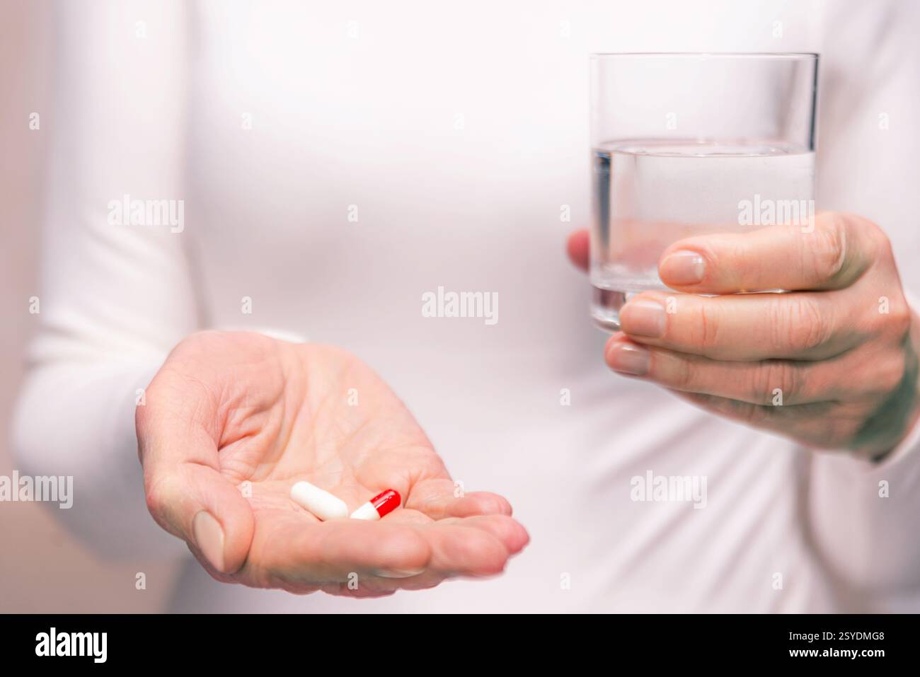 L'individuo si prepara a prendere il farmaco tenendo le capsule in una mano e un bicchiere d'acqua nell'altra mentre è in piedi all'interno. Foto Stock