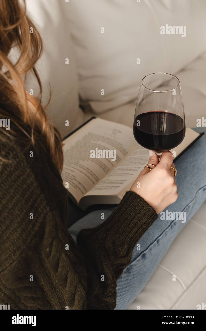 Una persona con lunghi capelli ondulati che indossa un maglione verde scuro si siede comodamente, reggendo un bicchiere di vino rosso mentre legge un libro su un divano grigio chiaro. Foto Stock