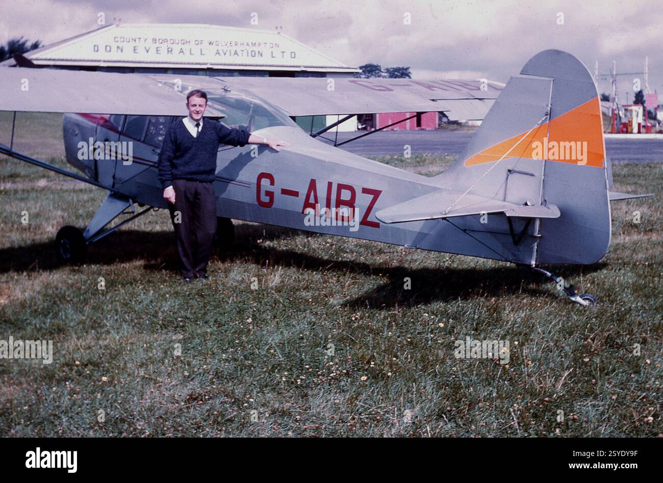 1969, un uomo in piedi accanto a un aereo leggero - un Auster J/1N Alpha G-AIBZ 2161 - sull'erba dell'aeroporto di Pendeford a Wolverhampton, Inghilterra, Regno Unito. Le officine e l'appendiabiti di Don Everall Aviation Ltd sullo sfondo. In questo periodo, l'operatore locale di pullman Don Everall aveva una divisione aerea che aveva sede a Pendeford e all'aeroporto di Elmdon, in seguito chiamato aeroporto internazionale di Birmingham. L'attività di trasporto di Don Everall nelle grandi midlands risale agli anni '1920, quando Don e suo fratello acquistarono il primo pullman a Wolverhampton. Foto Stock