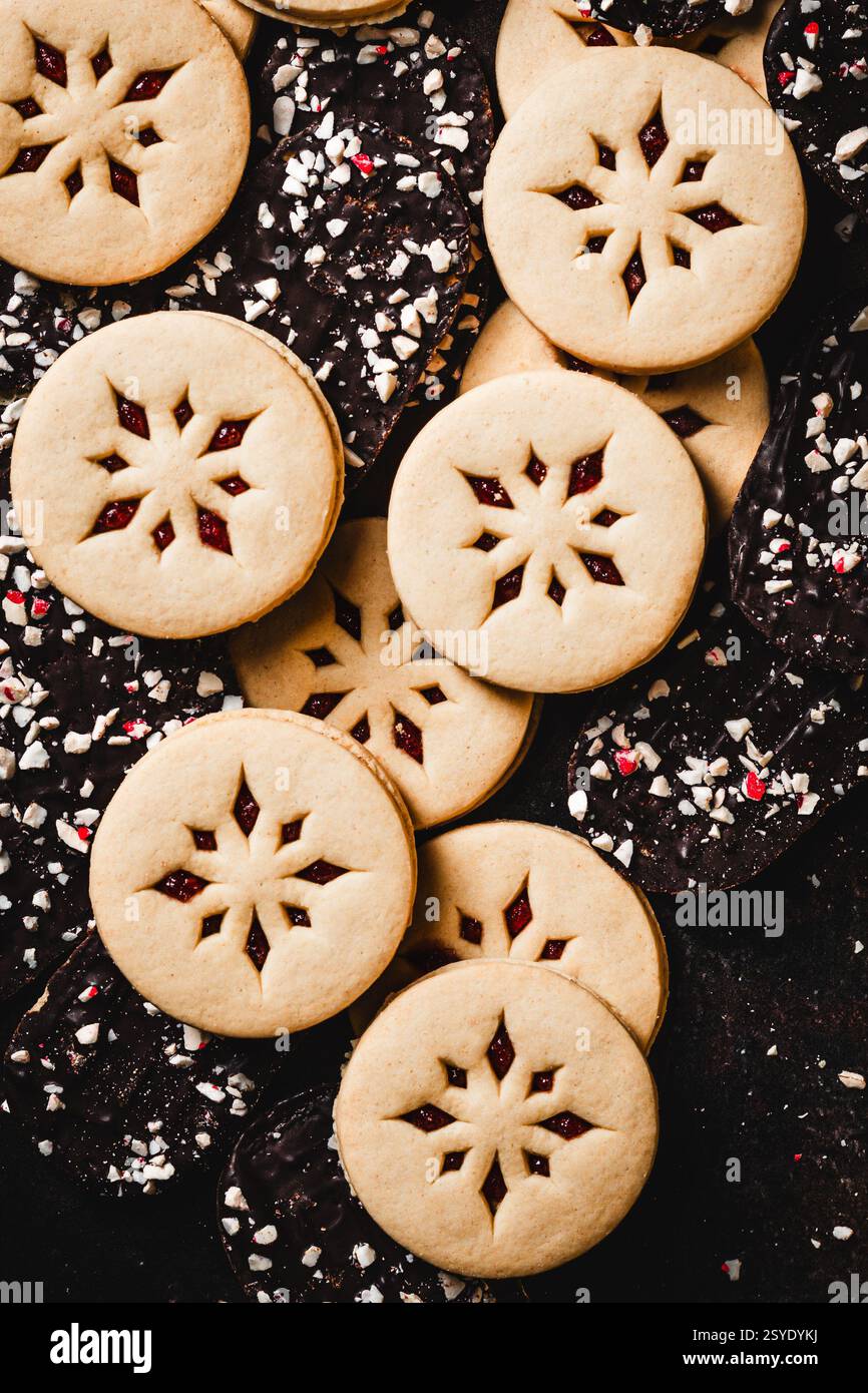 Un assortimento festoso di biscotti Linzer con fiocchi di neve con marmellata di lamponi e corteccia di menta piperita al cioccolato fondente, creando un elegante dessert natalizio. Foto Stock