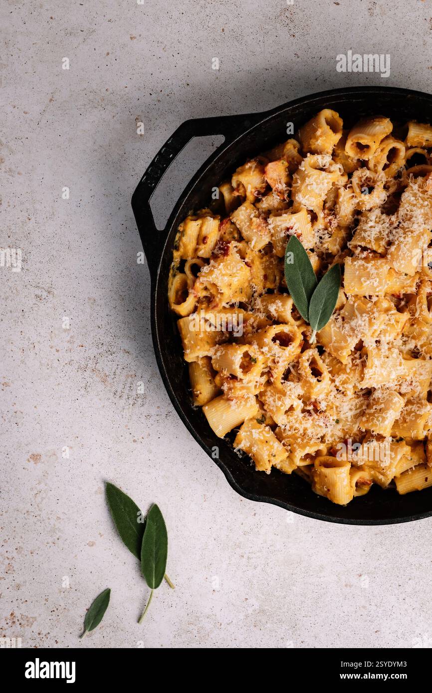 Una padella rustica in ghisa ripiena di cremosa pasta rigatoni, pancetta croccante e parmigiano, guarnita con salvia fresca su un backgroun neutro Foto Stock