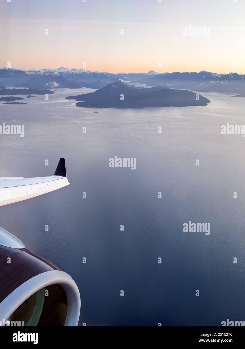 Una vista dalla finestra di un aereo che si affaccia sulle isole costiere e sulle montagne all'alba. L'ala dell'aereo e il motore contrastano con il vasto paesaggio marino sottostante. Foto Stock