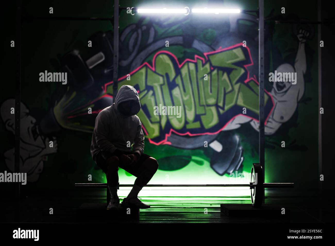 Un giovane atleta maschile siede solennemente accanto a un barbell in una palestra adornata da vibranti graffiti, sotto un'illuminazione soffusa, che riflette concentrazione e determinazione Foto Stock