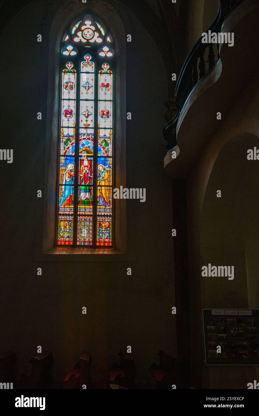 Una vibrante finestra di vetro colorato raffigurante figure e motivi colorati si illumina contro l'interno di un edificio con una scala curva poco illuminata Foto Stock