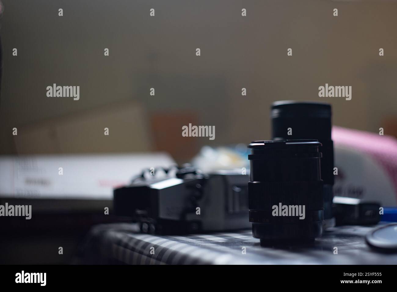 Varie attrezzature fotografiche sono sparse su una tovaglia a scacchi in una stanza delicatamente illuminata. Gli obiettivi e il corpo della fotocamera riflettono la luce ambientale, creando un Foto Stock