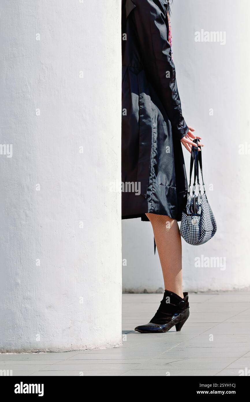 Una donna elegante che indossa un trench nero, calze a rete e stivaletti, tenendo in mano una borsa pied de poule accanto a una colonna bianca. Foto Stock