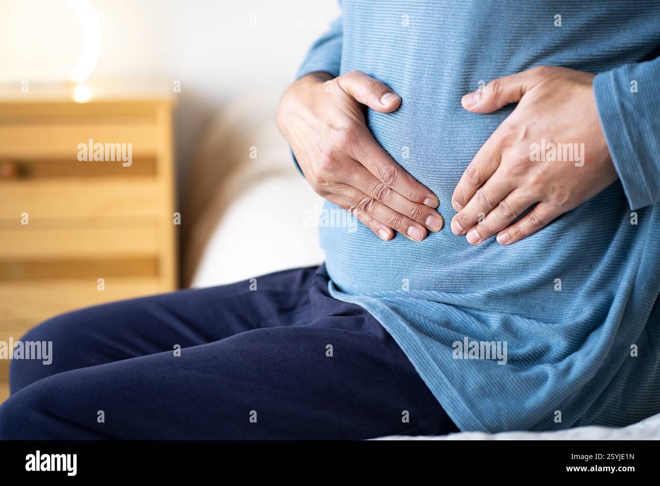 Uomo seduto da solo sul suo letto e soffre di crampi allo stomaco mentre era a casa da solo. Il dolore addominale può essere causato da problemi digestivi, infezioni, intest Foto Stock