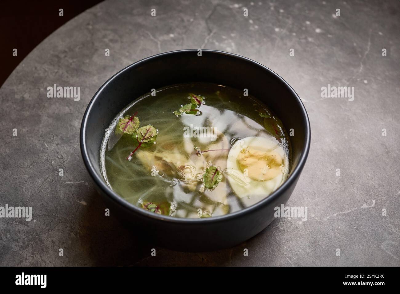 Zuppa limpida con uova, erbe e spaghetti nella ciotola nera su un tavolo in marmo grigio che ne esalta il fascino nutriente e confortevole, il concetto di stile gastronomico Foto Stock