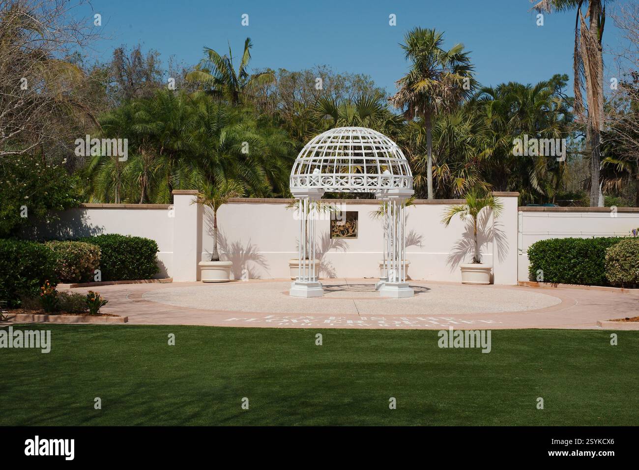 Gazebo alto bianco con giardino di ferro rotondo. Vista sull'erba verde con cespugli di arbusti verdi e palme laterali. Giorno luminoso e soleggiato con cielo azzurro. W Foto Stock