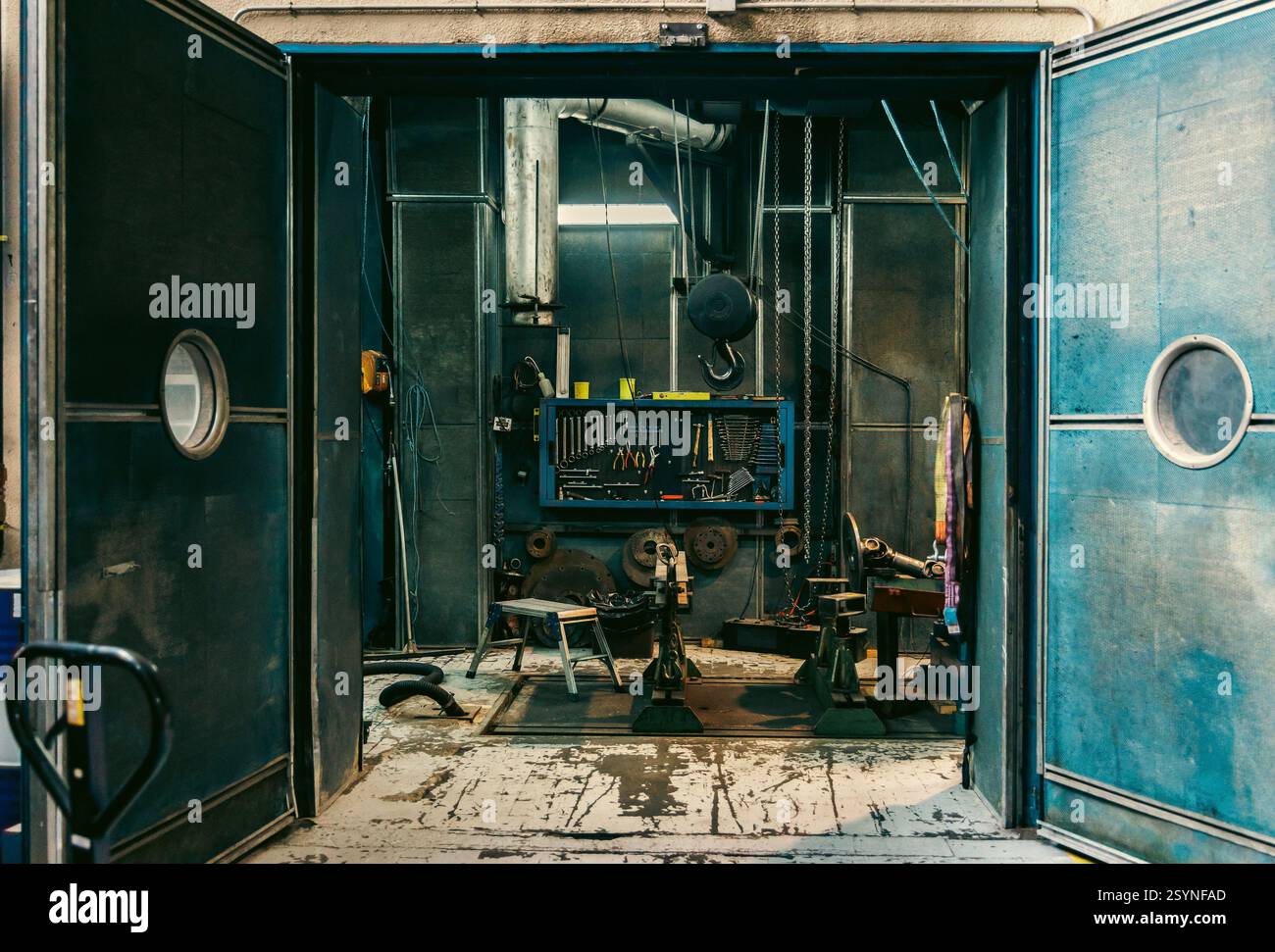 Grandi porte industriali blu si aprono per rivelare uno spazio interno con apparecchiature, cavi e strutture metalliche in fabbrica. Foto Stock