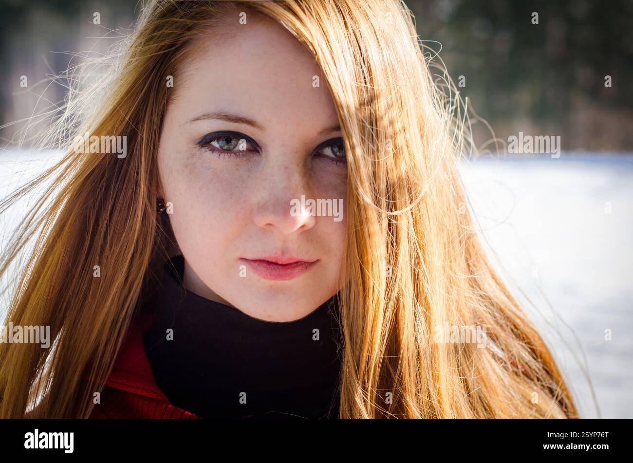 Giovane donna adulta con i capelli rossi accesi in piedi all'aperto. Lo sfondo mostra un paesaggio innevato con alberi sfocati. Il suo sguardo diretto e il suo sottile sorriso co Foto Stock