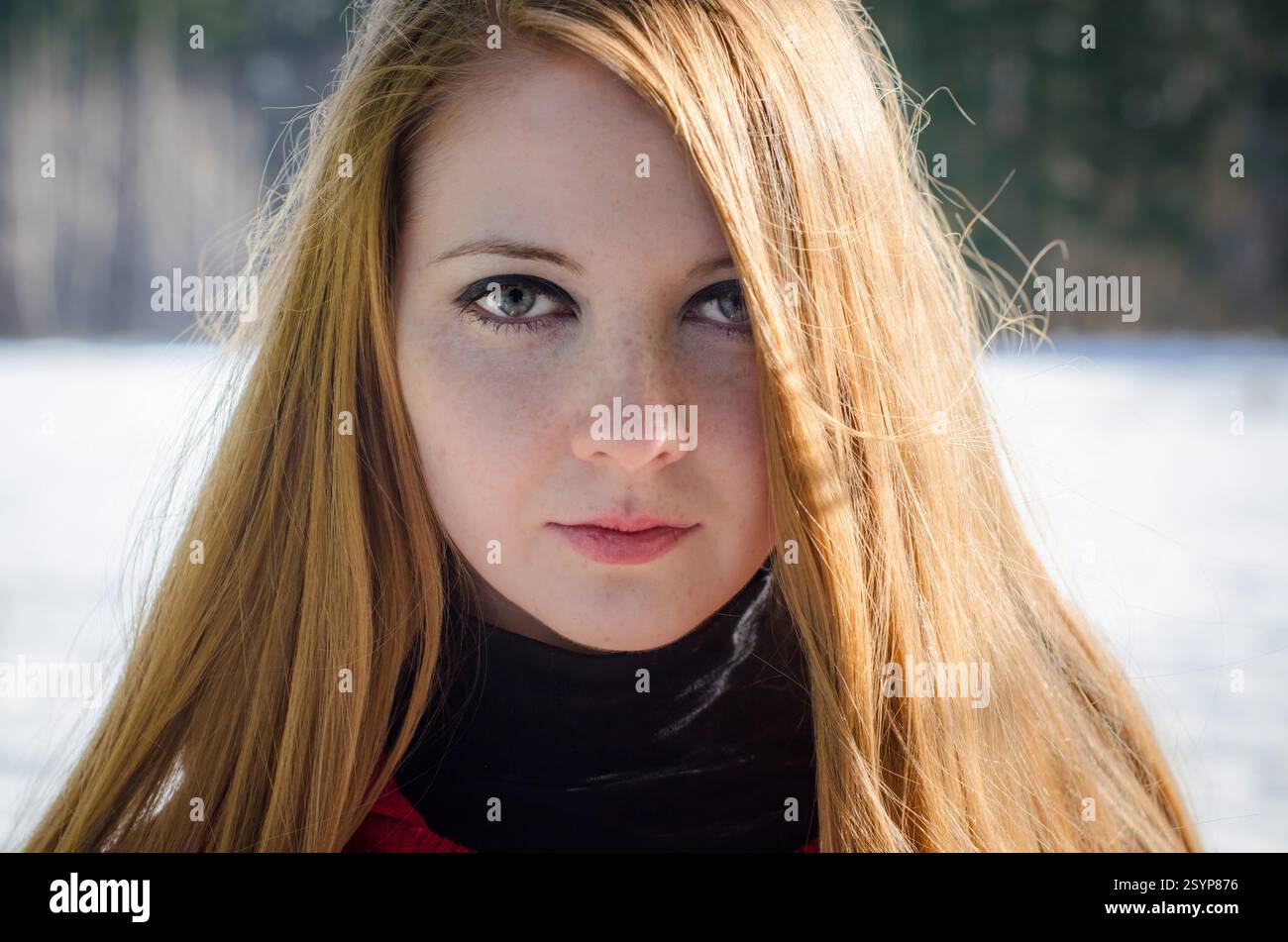 Giovane donna adulta con i capelli rossi accesi in piedi all'aperto. Lo sfondo mostra un paesaggio innevato con alberi sfocati. Il suo sguardo diretto e il suo sottile sorriso co Foto Stock