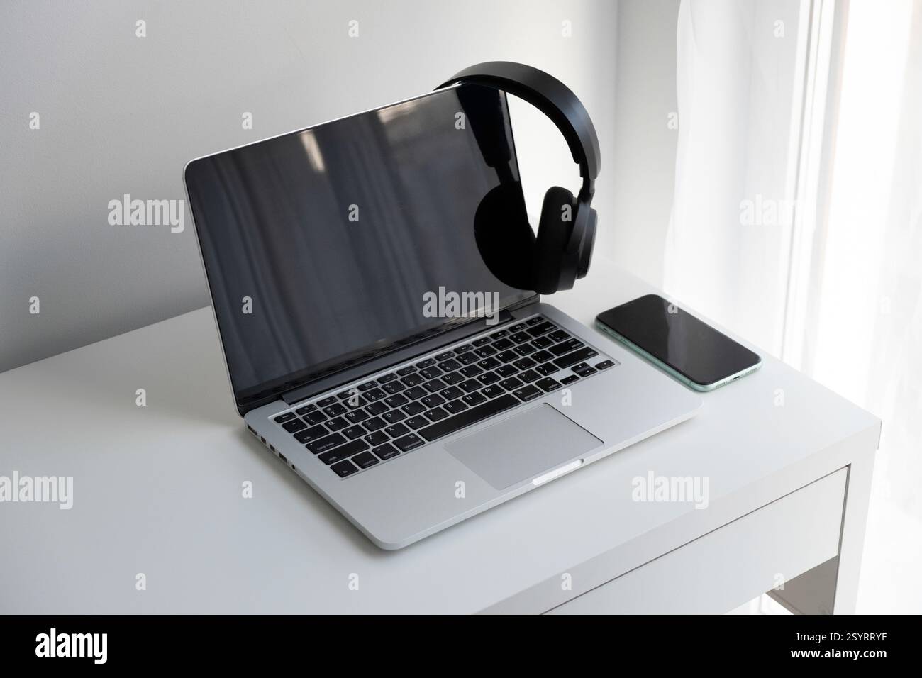 Laptop argento aperto, cuffie wireless nere e smartphone su una scrivania bianca Foto Stock
