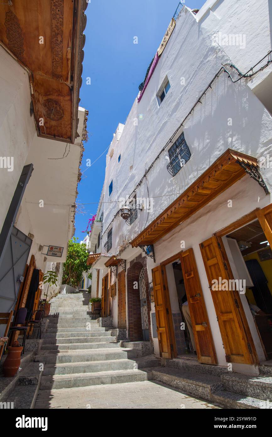 Strada stretta con edifici imbiancati, porte e persiane in legno e una scala che conduce verso l'alto sotto un cielo azzurro, creando una me pittoresca Foto Stock
