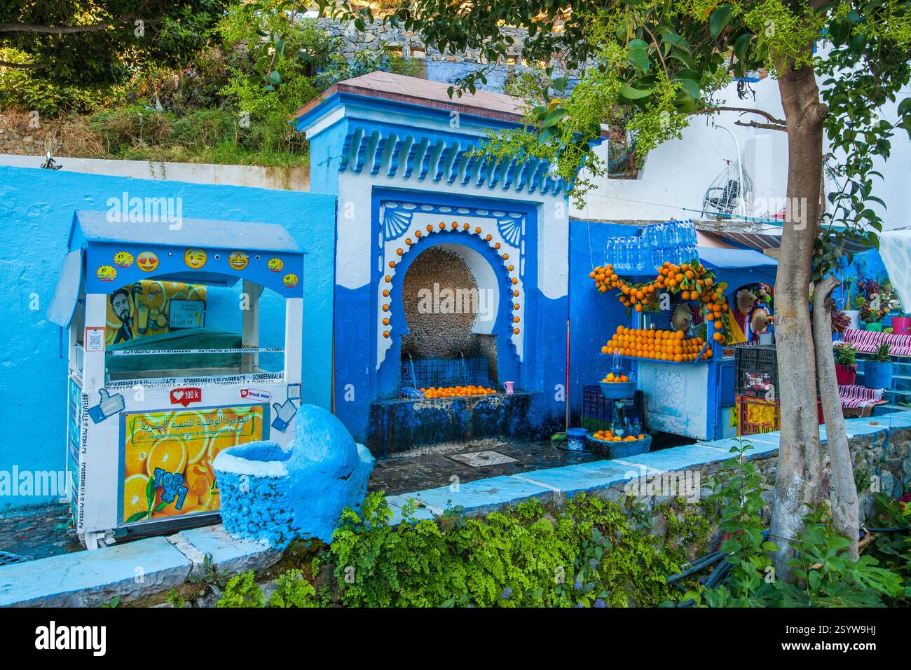scena vivace in una città dipinta di blu, caratterizzata da una fontana decorativa, un banco di frutta traboccante di arance e vegetazione lussureggiante, che crea un affascinante a Foto Stock