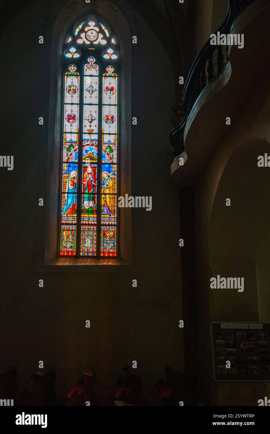 Una vibrante finestra di vetro colorato raffigurante figure e motivi colorati si illumina contro l'interno di un edificio con una scala curva poco illuminata Foto Stock