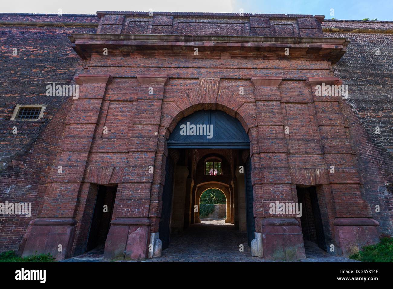 Un grande cancello ad arco in mattoni con una porta in metallo scuro e un passaggio che conduce attraverso, visto direttamente di fronte in una giornata di sole. Foto Stock