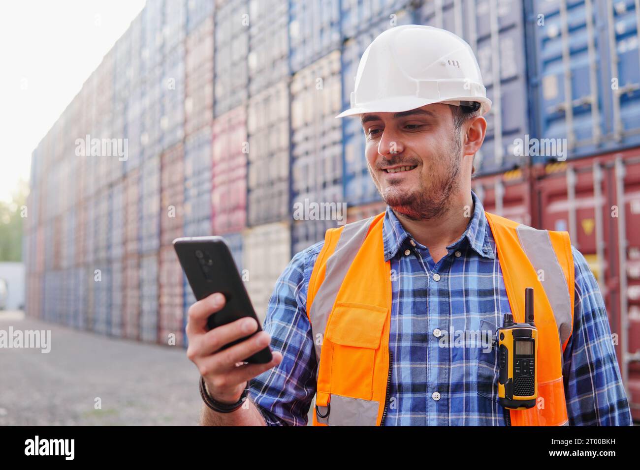 Giovane ingegnere nel casco che usa il telefono nel magazzino container. Gestione logistica nel deposito container. Foto Stock