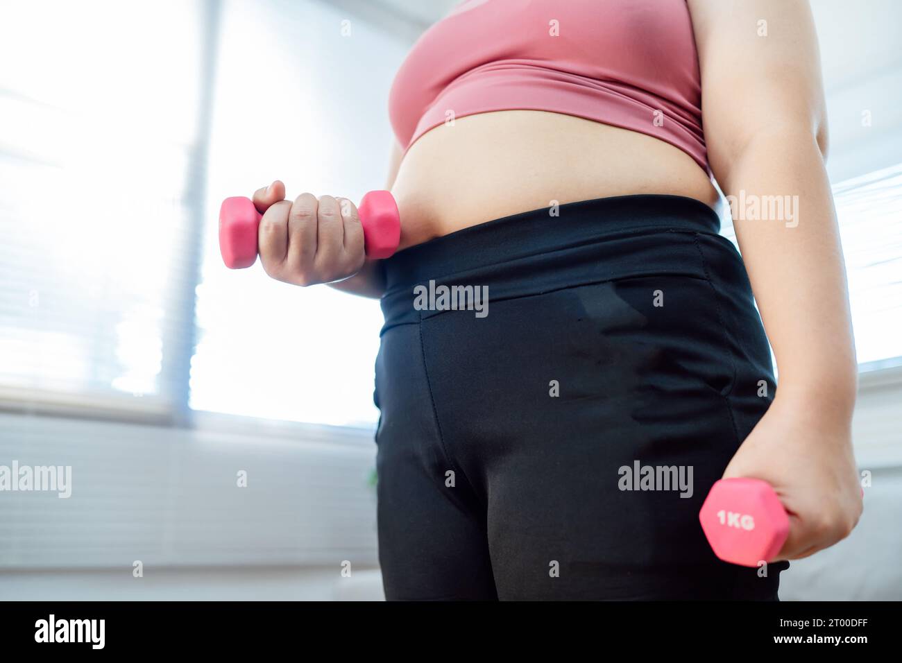 Donna in sovrappeso che si esercita per perdita di peso. Allenati con i manubri in posizioni strette a casa tua in salotto allegramente Foto Stock