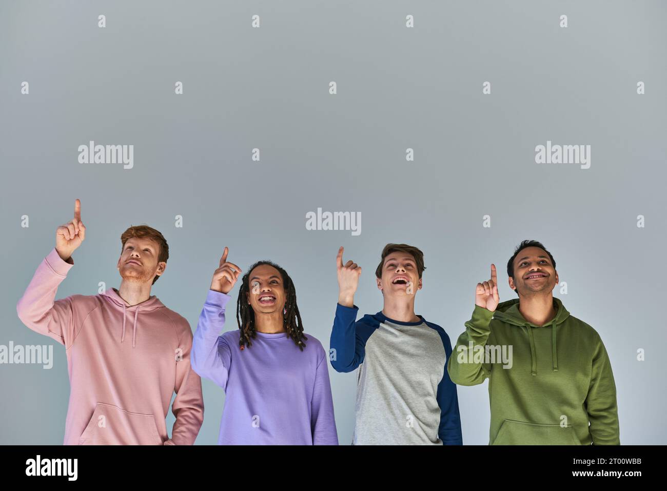 quattro uomini gioiosi in strada indossano lo sguardo verso l'alto e puntano verso l'alto su sfondo grigio, diversità culturale Foto Stock