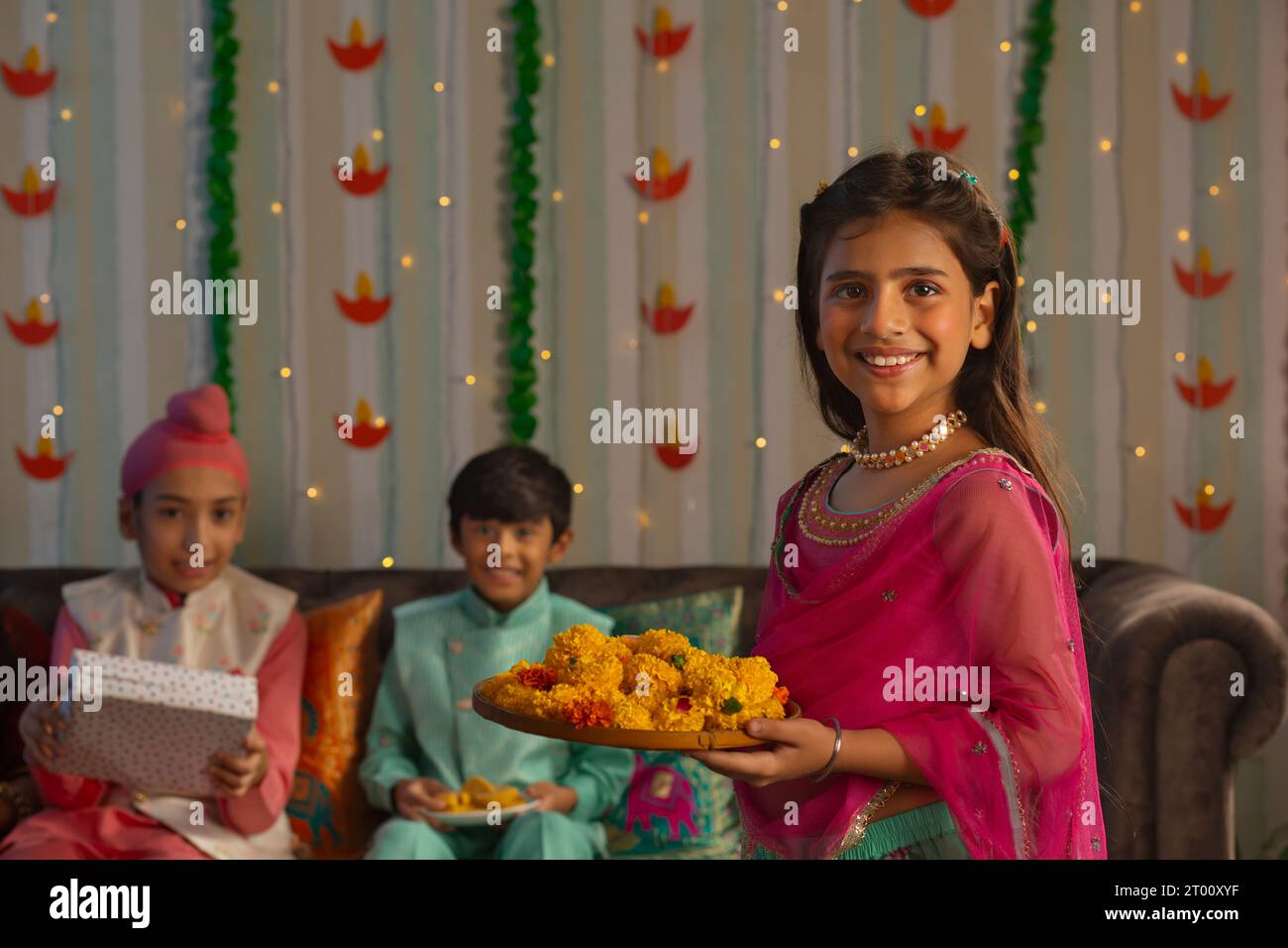 Ragazza allegra in piedi con un piatto di fiori in occasione di Diwali e dei suoi amici seduti dietro Foto Stock
