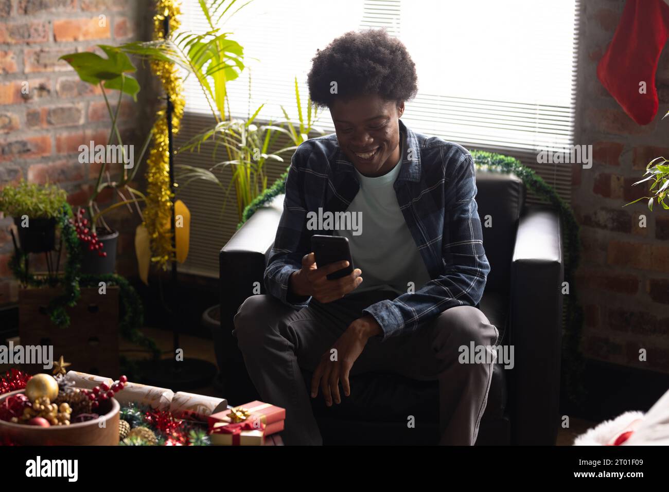 Felice uomo afro-americano che usa lo smartphone seduto in poltrona a casa durante il periodo natalizio Foto Stock