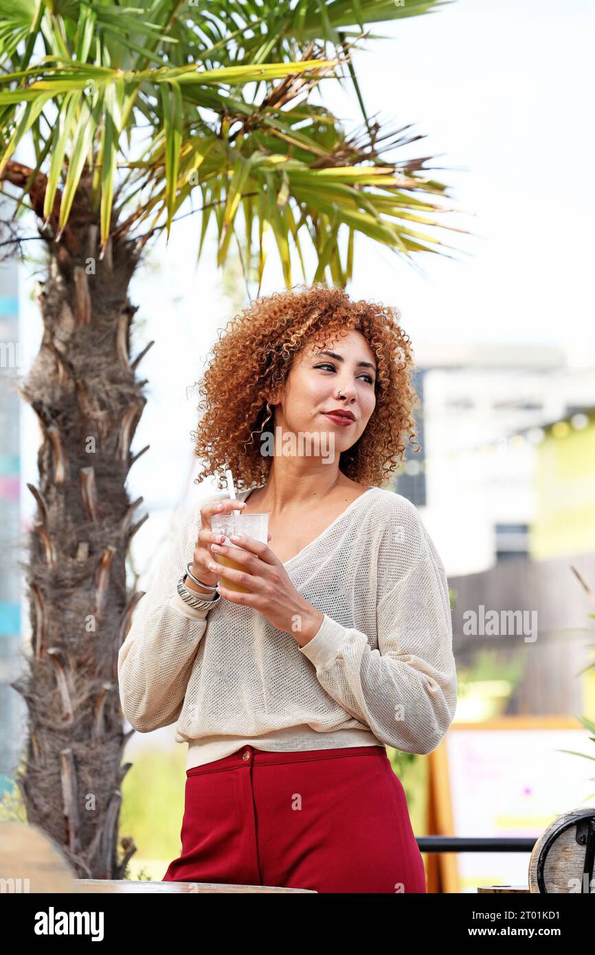 Donna dai capelli ricci con maglione bianco e pantaloni rossi che si diverte all'aperto sotto un albero di palmer in un paese tropicale Foto Stock