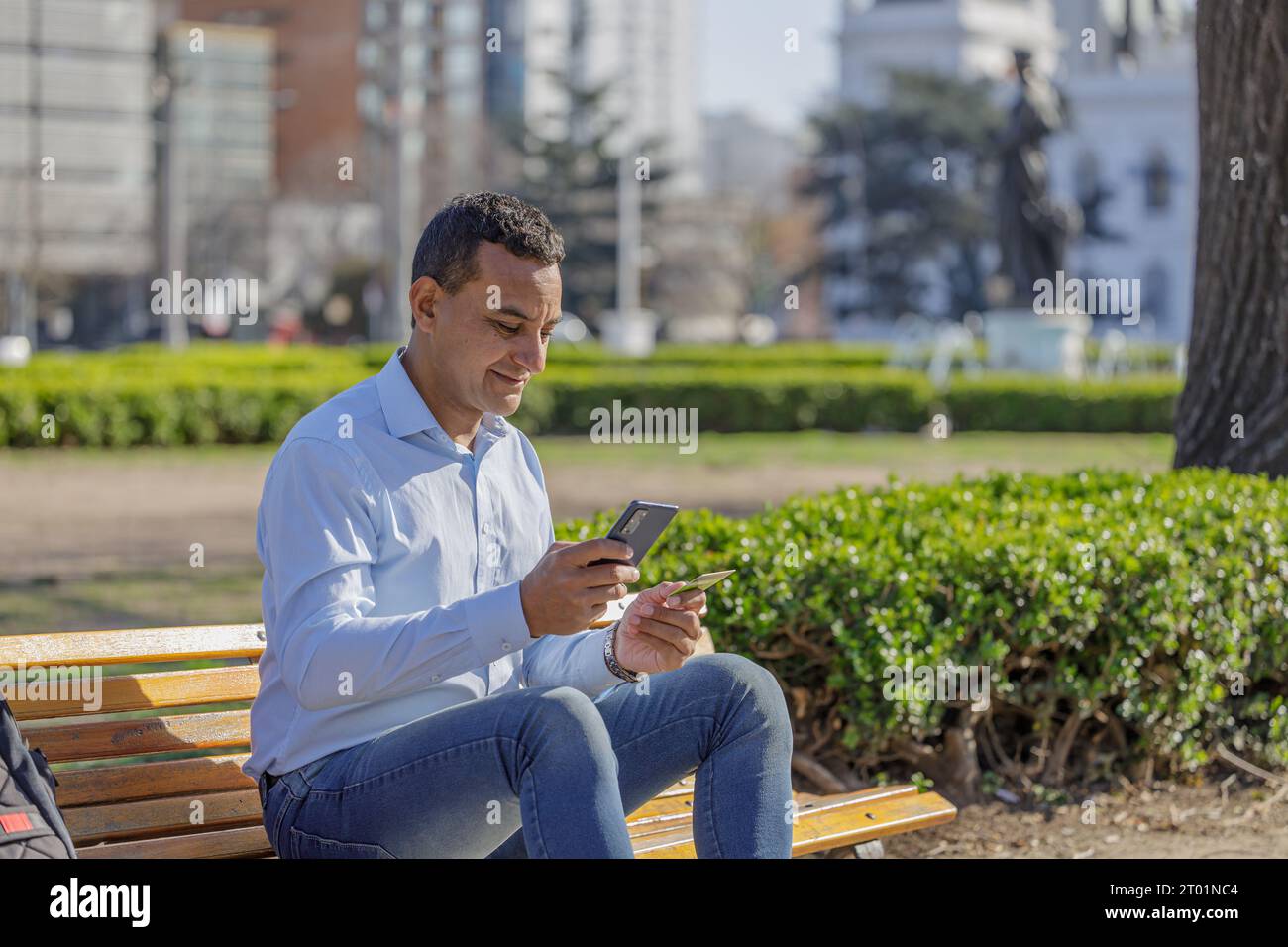 Un uomo latino che fa un acquisto con una carta di credito sul suo telefono cellulare in una panchina quadrata. Foto Stock