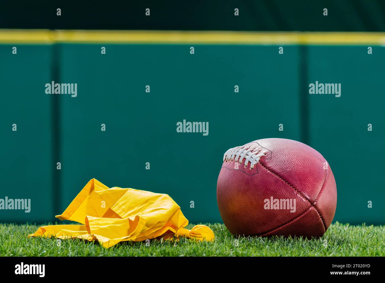 Una vista dall'angolo basso di una bandiera di rigore gialla lanciata da un arbitro accanto a un football americano professionista con parete imbottita verde sullo sfondo. Foto Stock