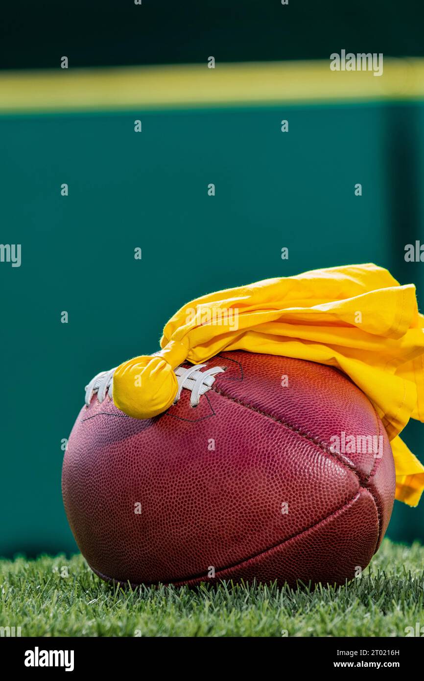 Una vista dall'angolo basso di una bandiera di rigore gialla lanciata da un arbitro accanto a un football americano professionista con parete imbottita verde sullo sfondo. Foto Stock