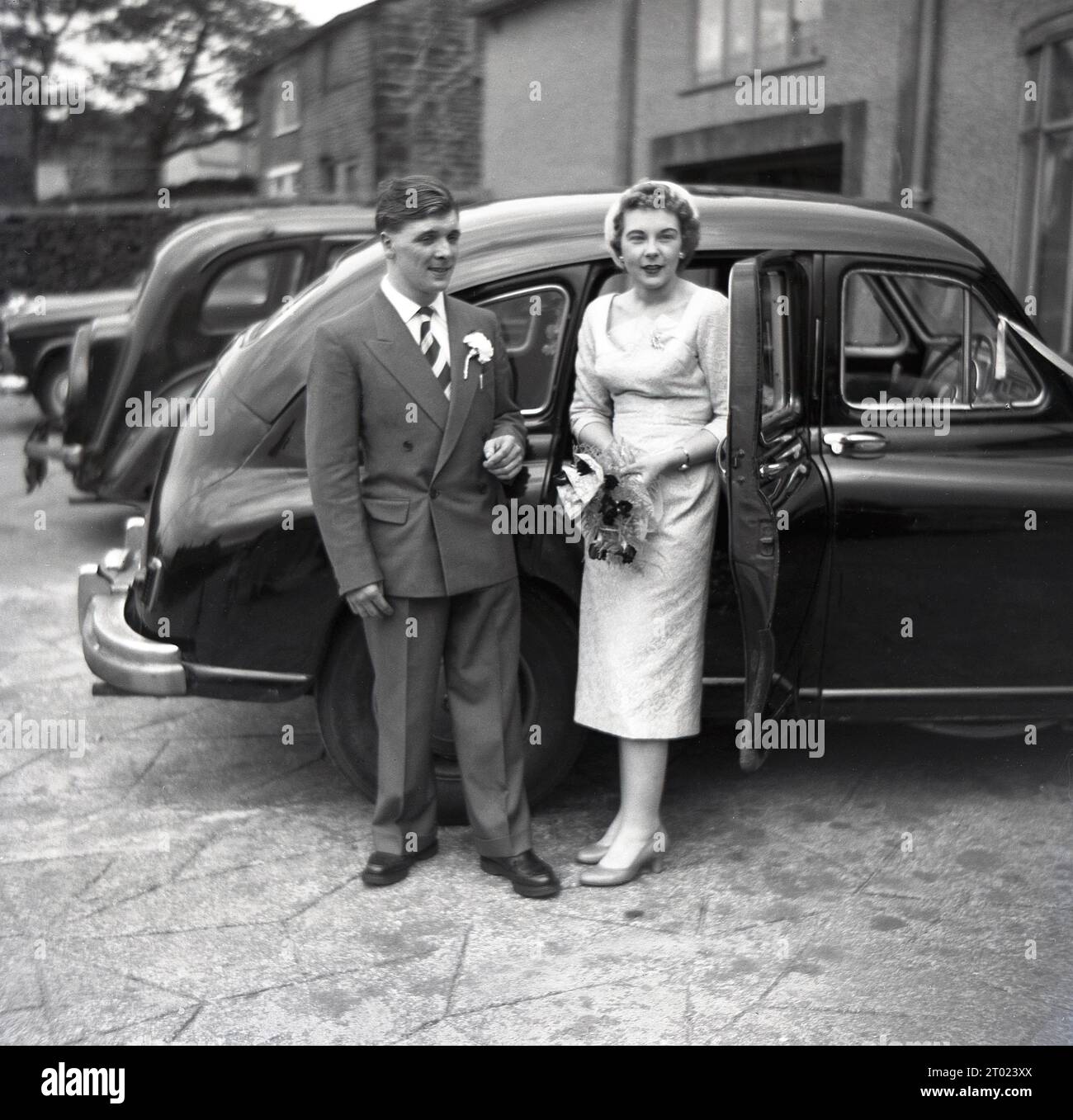 Anni '1960, storica, una giovane coppia appena sposata in piedi fuori dalla loro auto dell'epoca, la giovane signora che regge un bouquet di fiori, Inghilterra, Regno Unito. Foto Stock