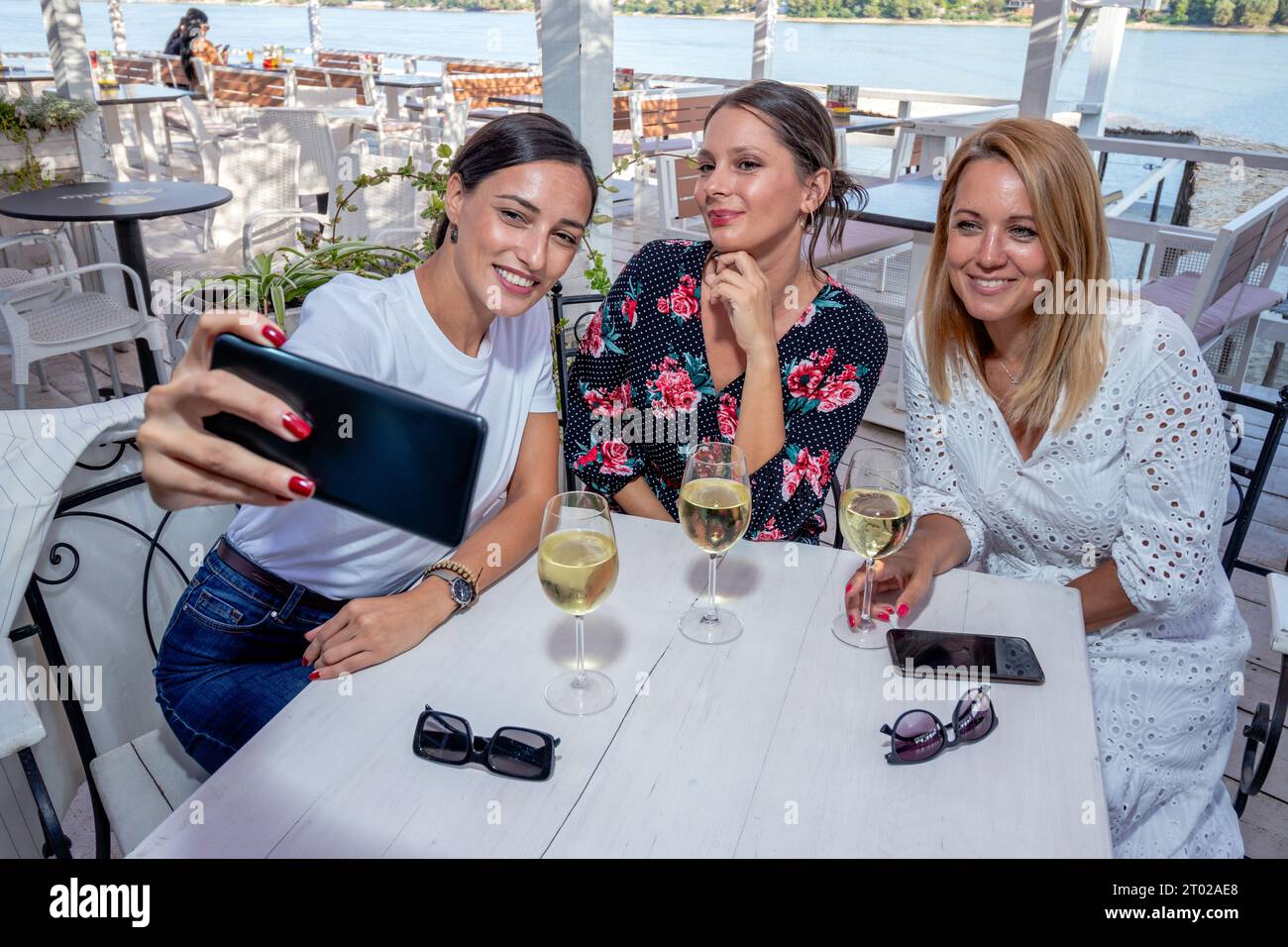 Tre amiche allegri siedono a un tavolo con un bicchiere di vino bevendo, parlando e scattando selfie sulla terrazza di un ristorante sul fiume. Foto Stock